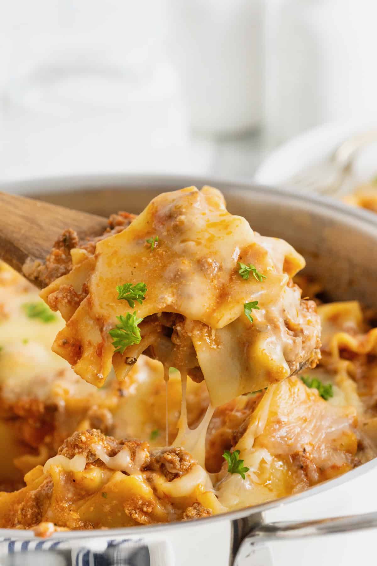 A wooden spoon scooping out some skillet lasagna from a stainless steel skillet.
