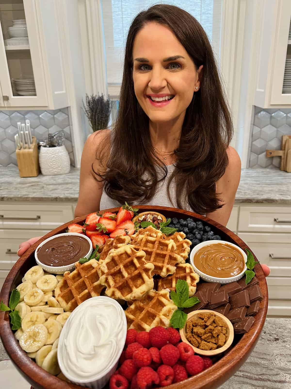 Cookbook author Maegan Brown wearing a sleeveless white top holding a large round wooden serving board filled with Belgian waffles, toppings, fresh fruit and whipped cream.