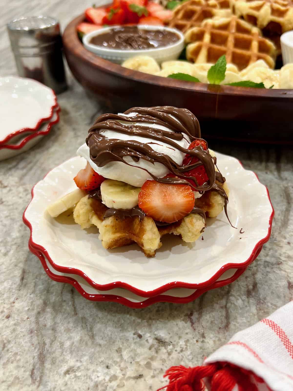 A belgian waffle piled high with banana slices, strawberry slices, whipped cream and Nutella on a red rimmed white plate.