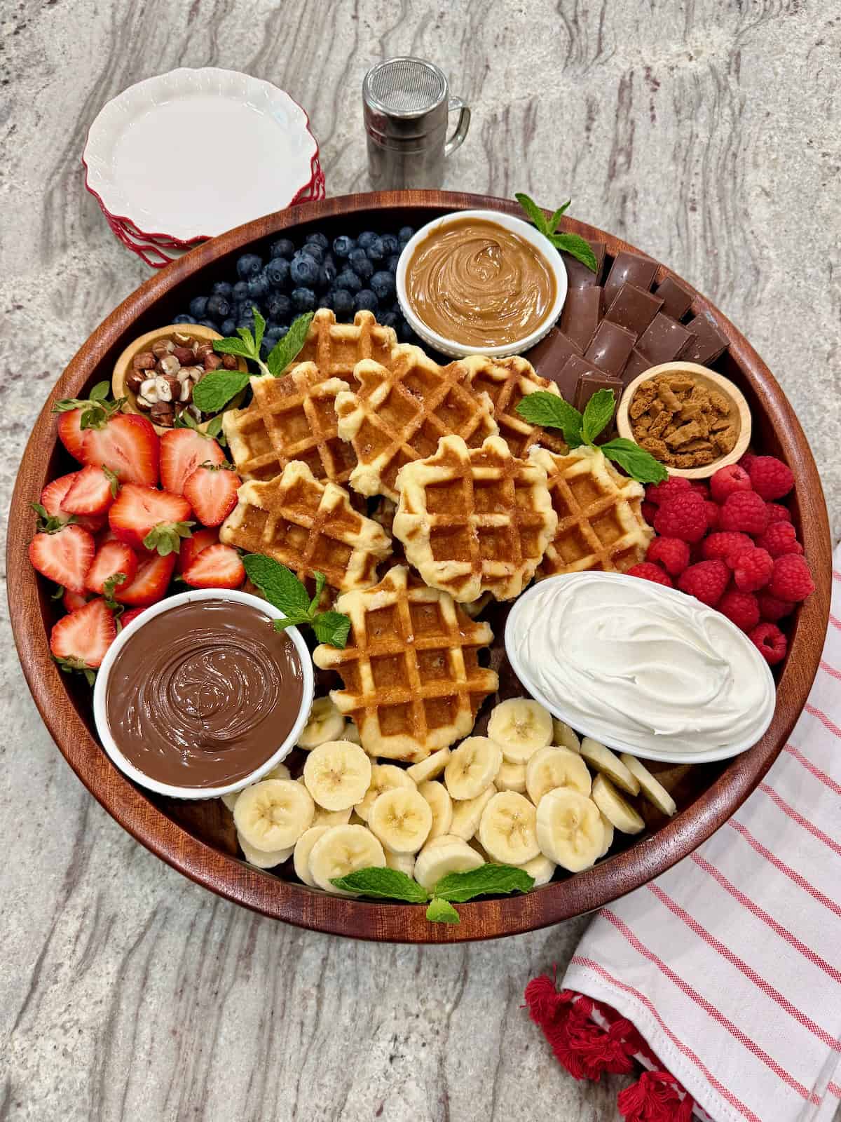 A round wood food tray loaded with Belgian waffles, fruit, Nutella, biscoff butter and whipped cream.