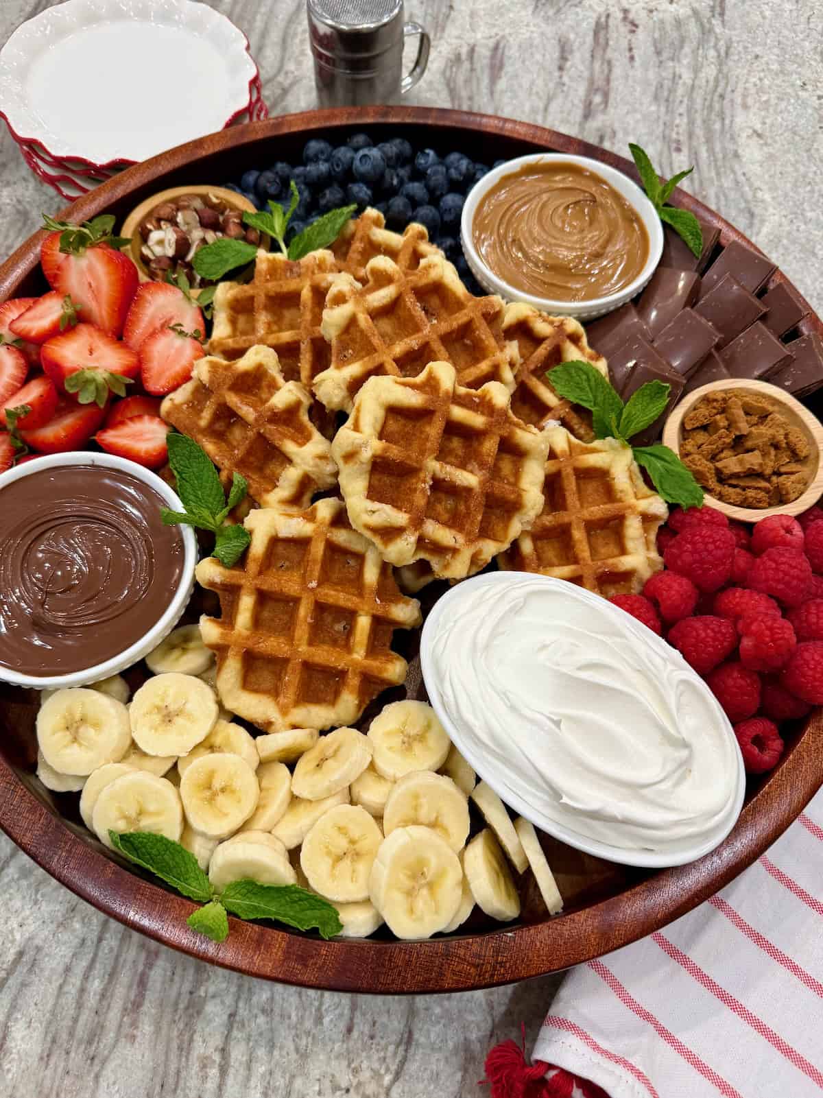 A round wood food tray loaded with Belgian waffles, fruit, Nutella, biscoff butter and whipped cream.