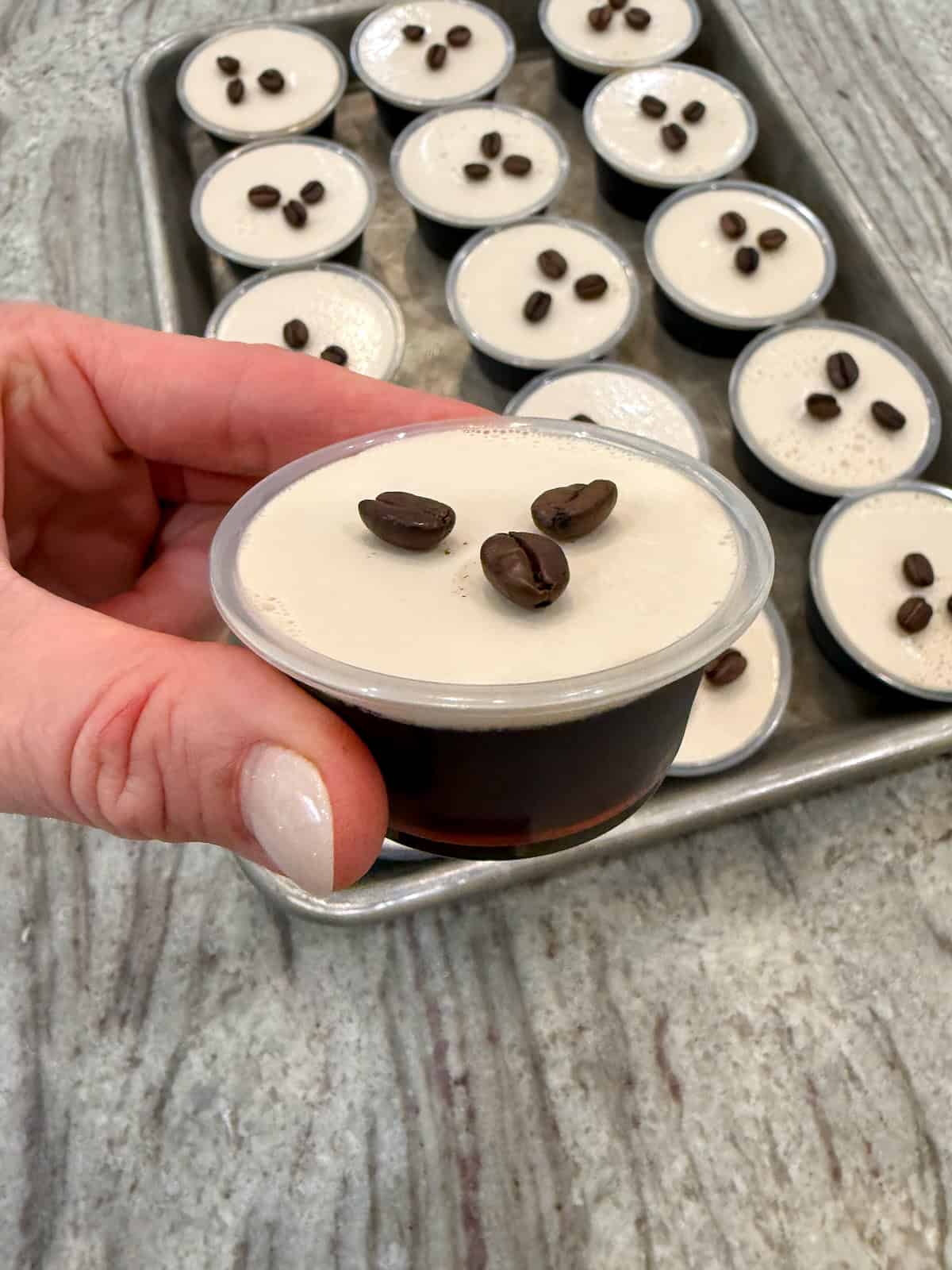 A hand holding an espresso martini Jell-O shot in front of a tray of shots.