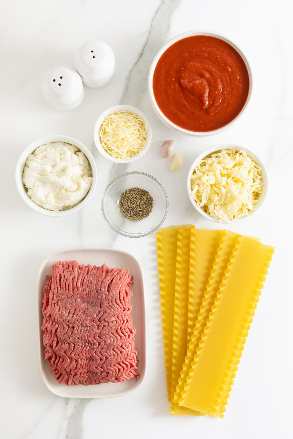 Ingredients to make cheesy skillet lasagna in small glass dishes on white marble counter.