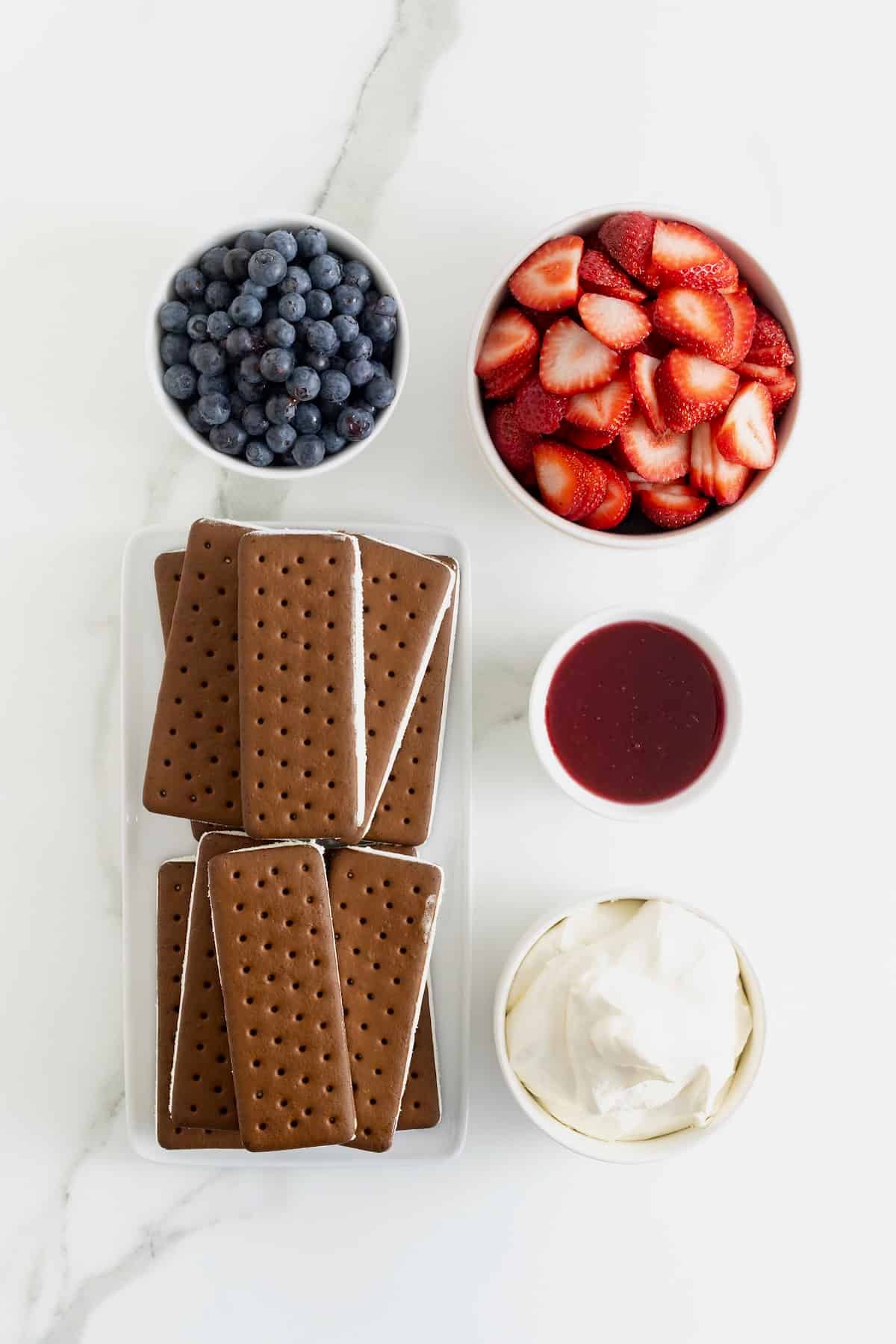 Ingredients to make a fresh berry ice cream sandwich cake in white dishes on a white marble counter.