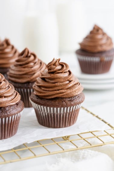 Five chocolate cupcakes with chocolate frosting on a parchment lined gold toned wire cooling rack.