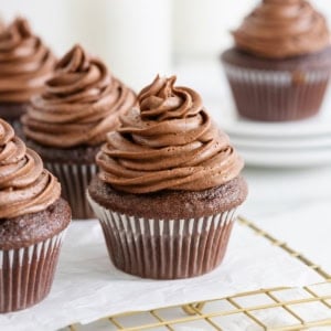 Five chocolate cupcakes with chocolate frosting on a parchment lined gold toned wire cooling rack.