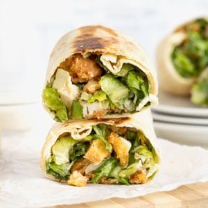 A crispy hot honey chicken caesar wrap cut in half on a parchment lined wood cutting board.