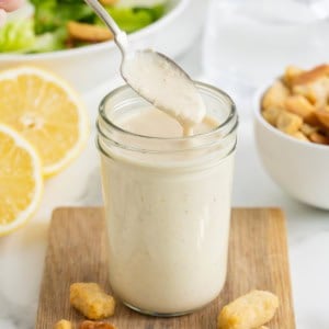 A spoon lifting caesar dressing out of a wide mouth jar o na small wood cutting board. There are halves of lemons in the background.