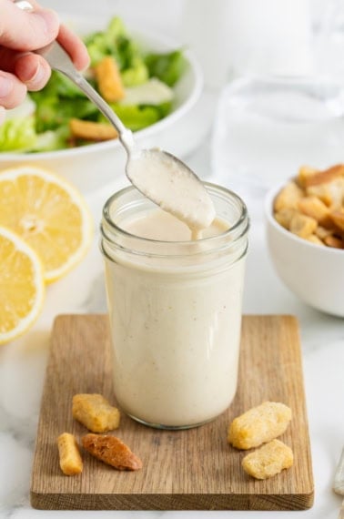 A spoon lifting caesar dressing out of a wide mouth jar o na small wood cutting board. There are halves of lemons in the background