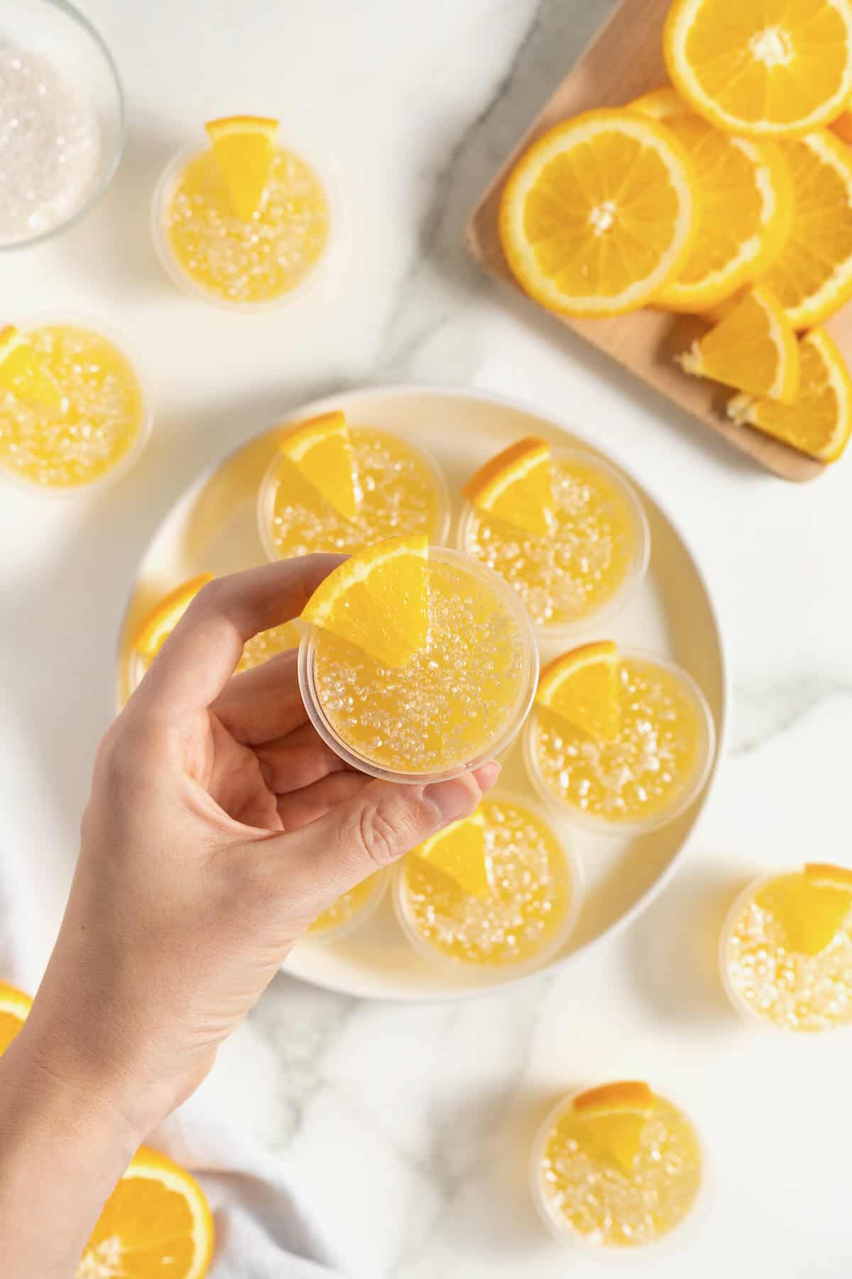 A hand holding a mimosa jello shot over a white platter of jello shots.