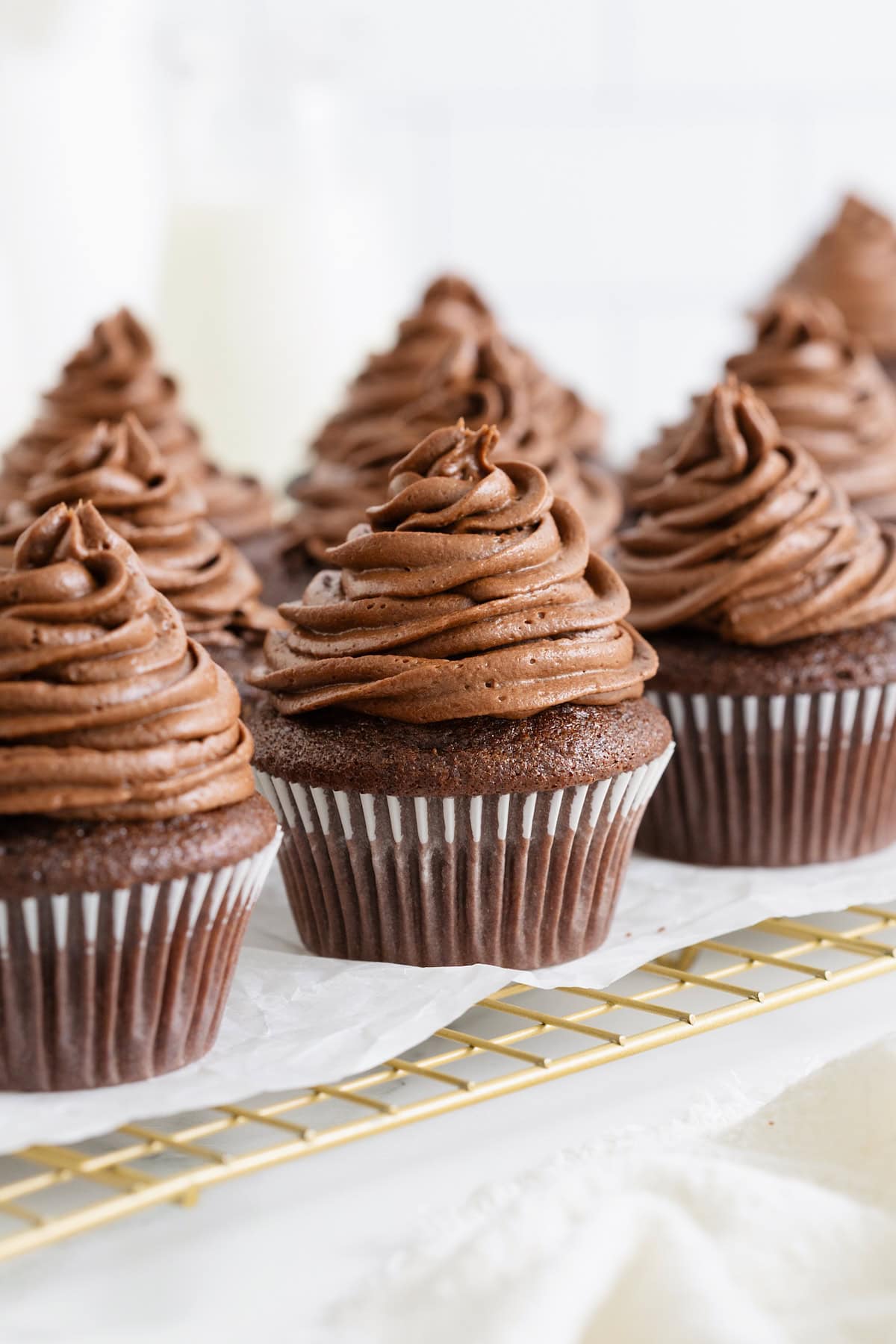 Six chocolate cupcakes with chocolate frosting on a parchment lined gold toned wire cooling rack.