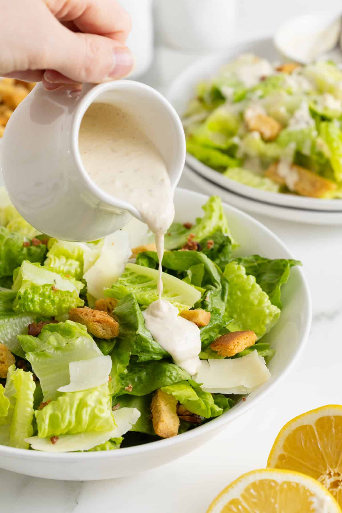 Caesar dressing being poured over a Caesar salad in a large white bowl.
