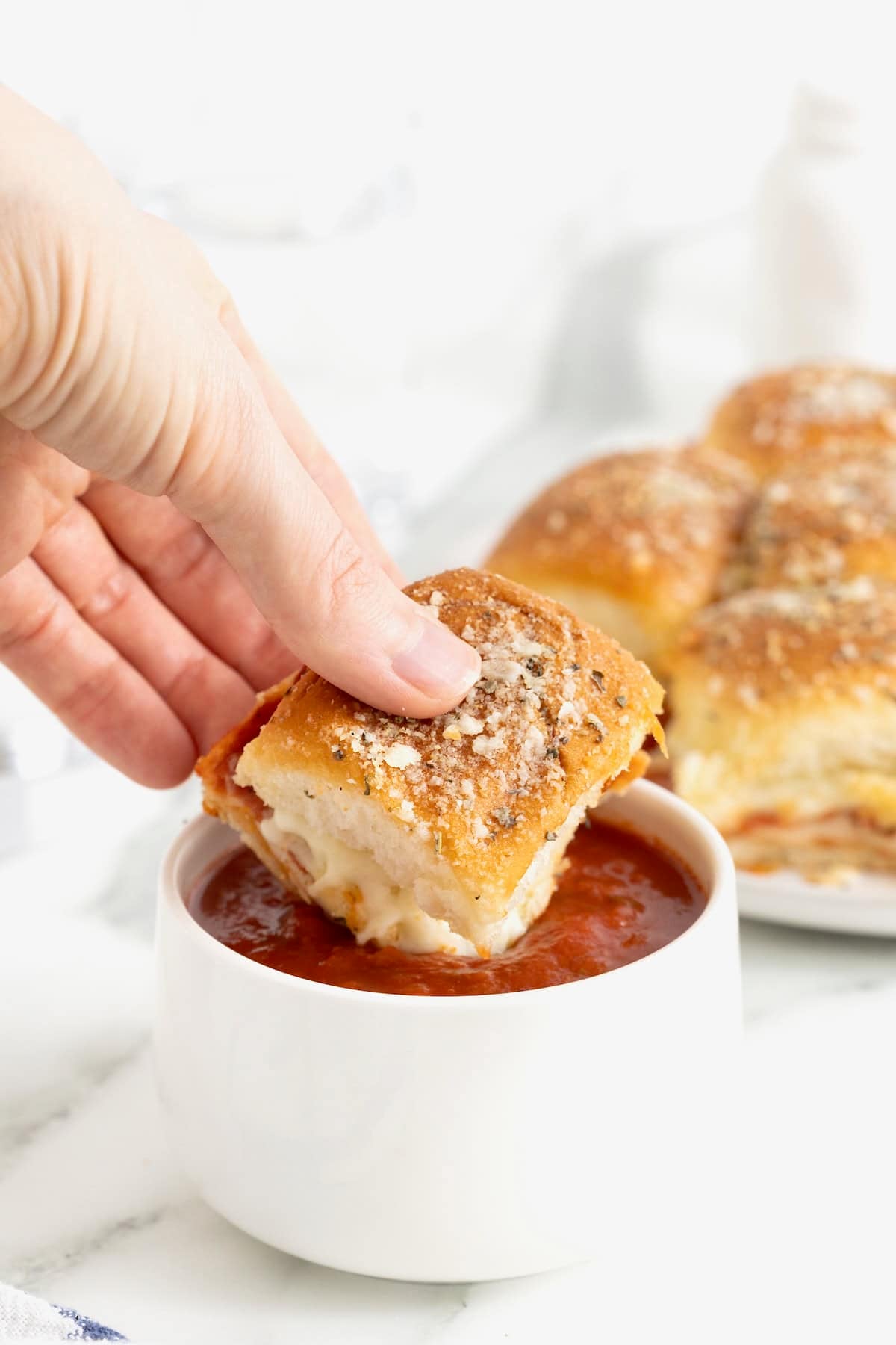 A hand holding a pizza slider dipping it into a small white dish of marinara sauce.