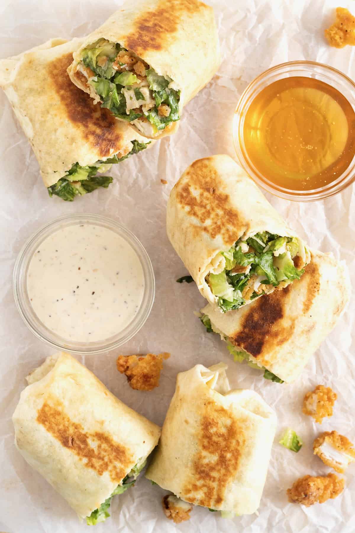 Chicken wraps scattered on a parchment lined cutting board next to small glass bows of honey and caesar dressing.