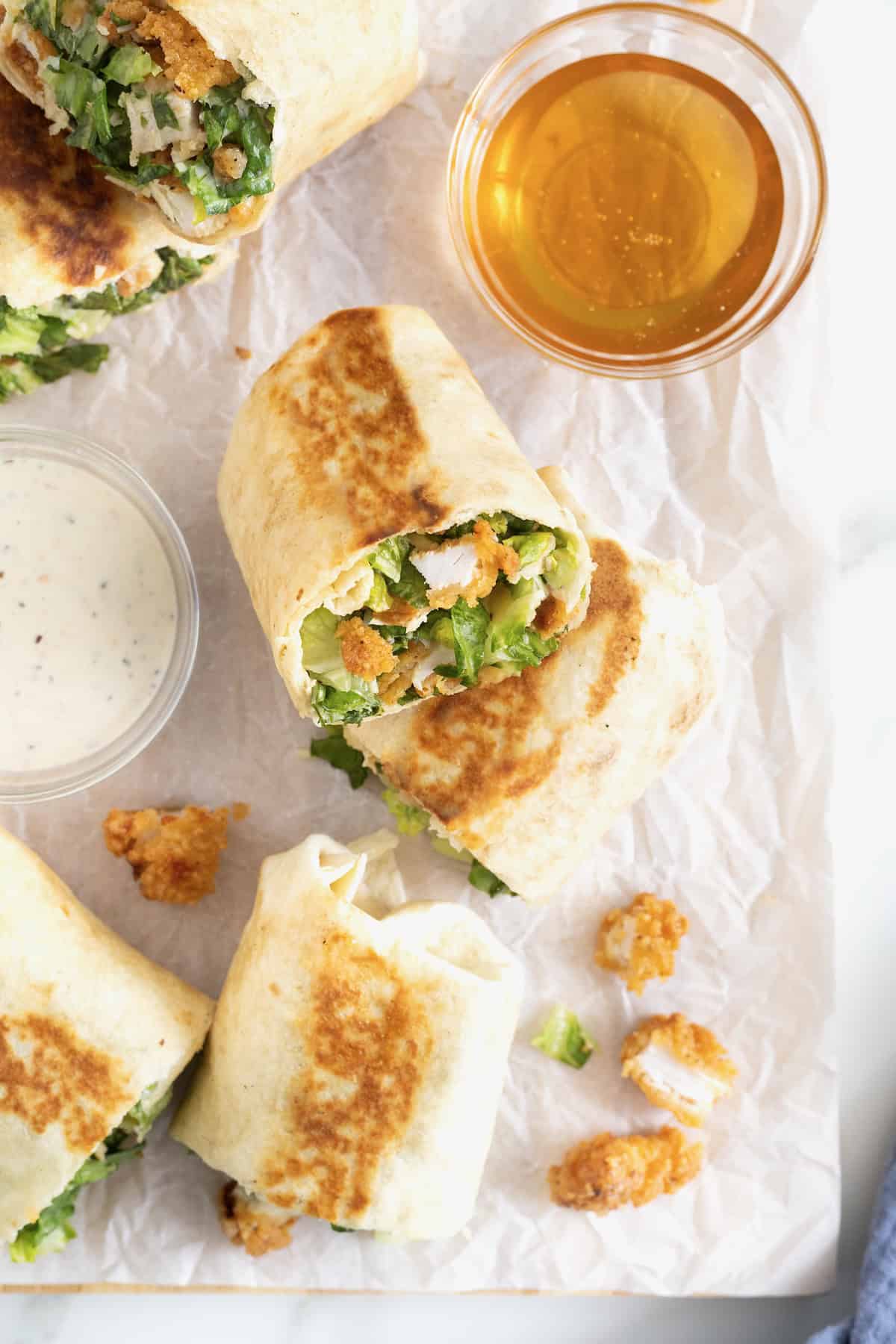 Chicken wraps scattered on a parchment lined cutting board next to small glass bows of honey and caesar dressing.