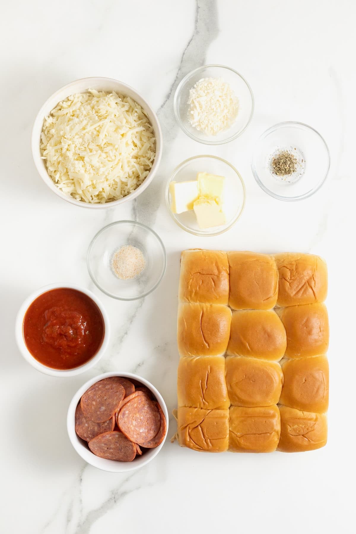 Ingredients to make pepperoni pizza sliders in small glass dishes on a white marble counter.