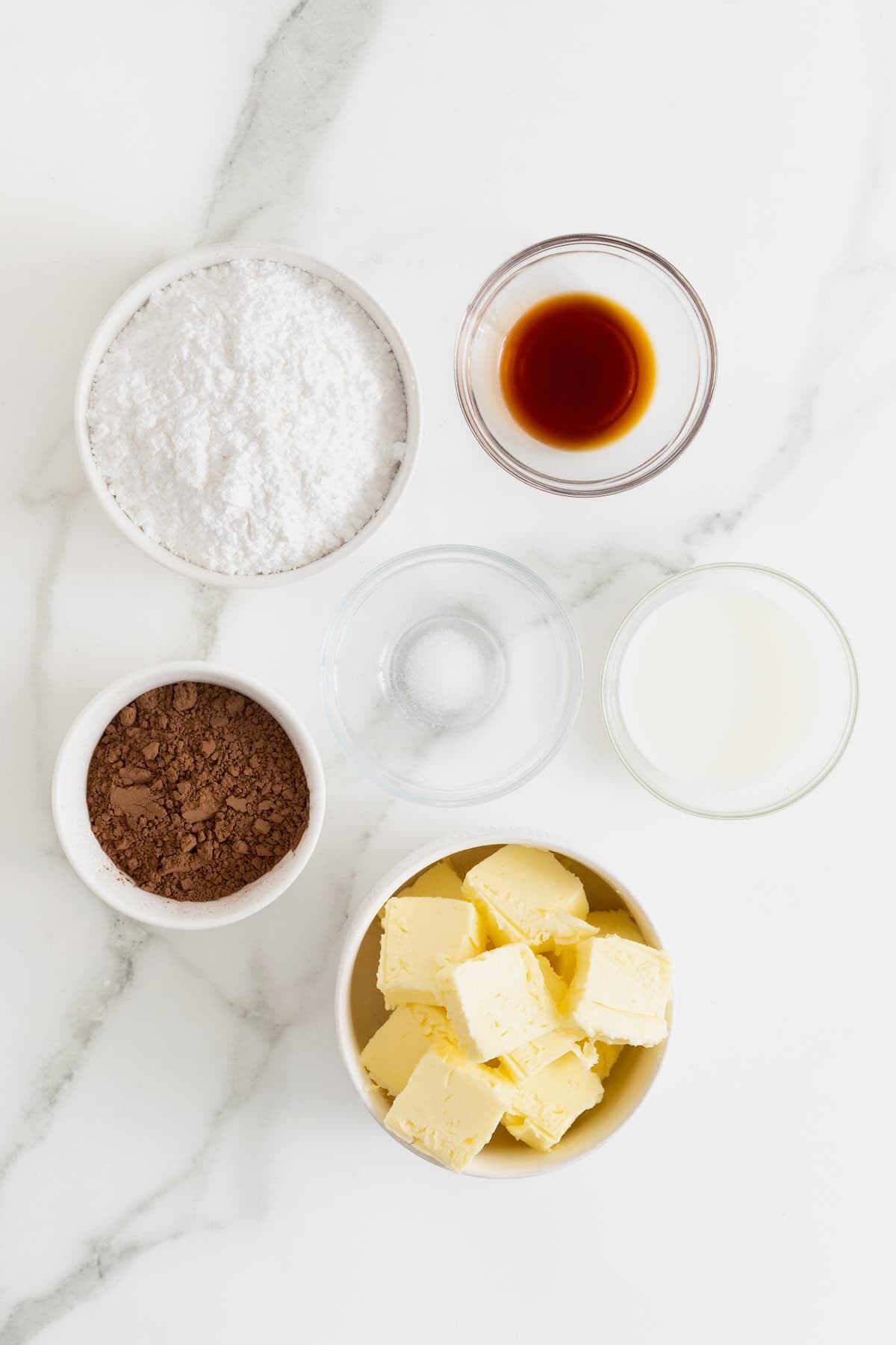 Ingredients to make chocolate buttercream frosting in small glass dishes on a white marble counter.