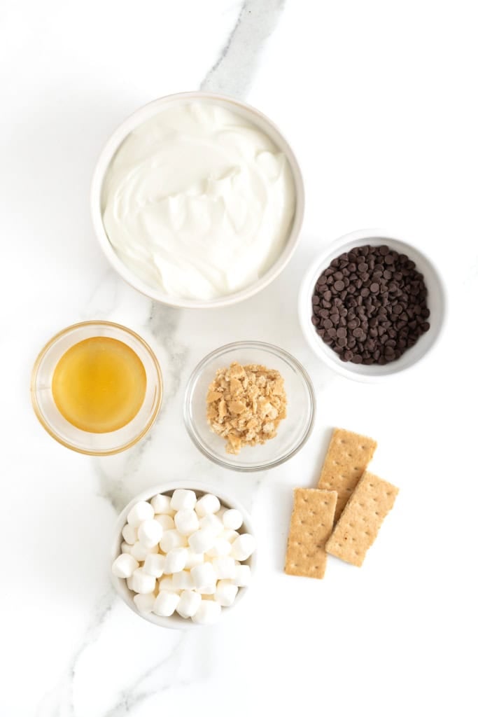 Ingredients to make frozen yogurt s'mores bars in small glass dishes on a white marble counter.