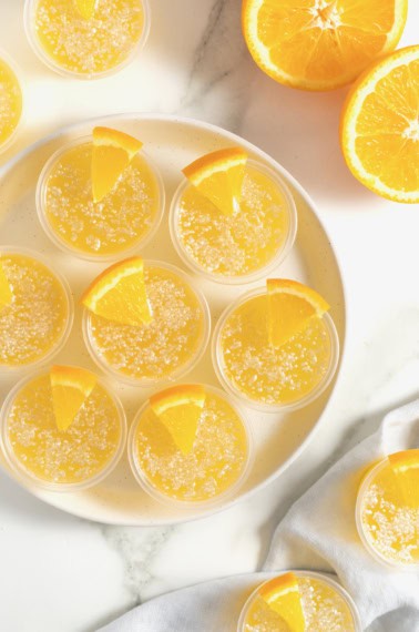 Seven mimosa jello shots in plastic shot cups rimmed in sugar and garnished with a small orange wedge.