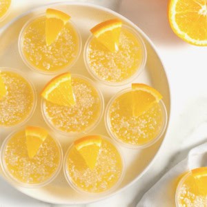 Seven mimosa jello shots in plastic shot cups rimmed in sugar and garnished with a small orange wedge.