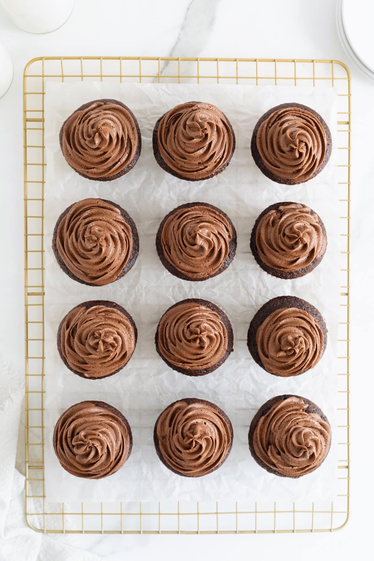 A dozen chocolate cupcakes with chocolate frosting on a parchment lined gold toned cooling rack.