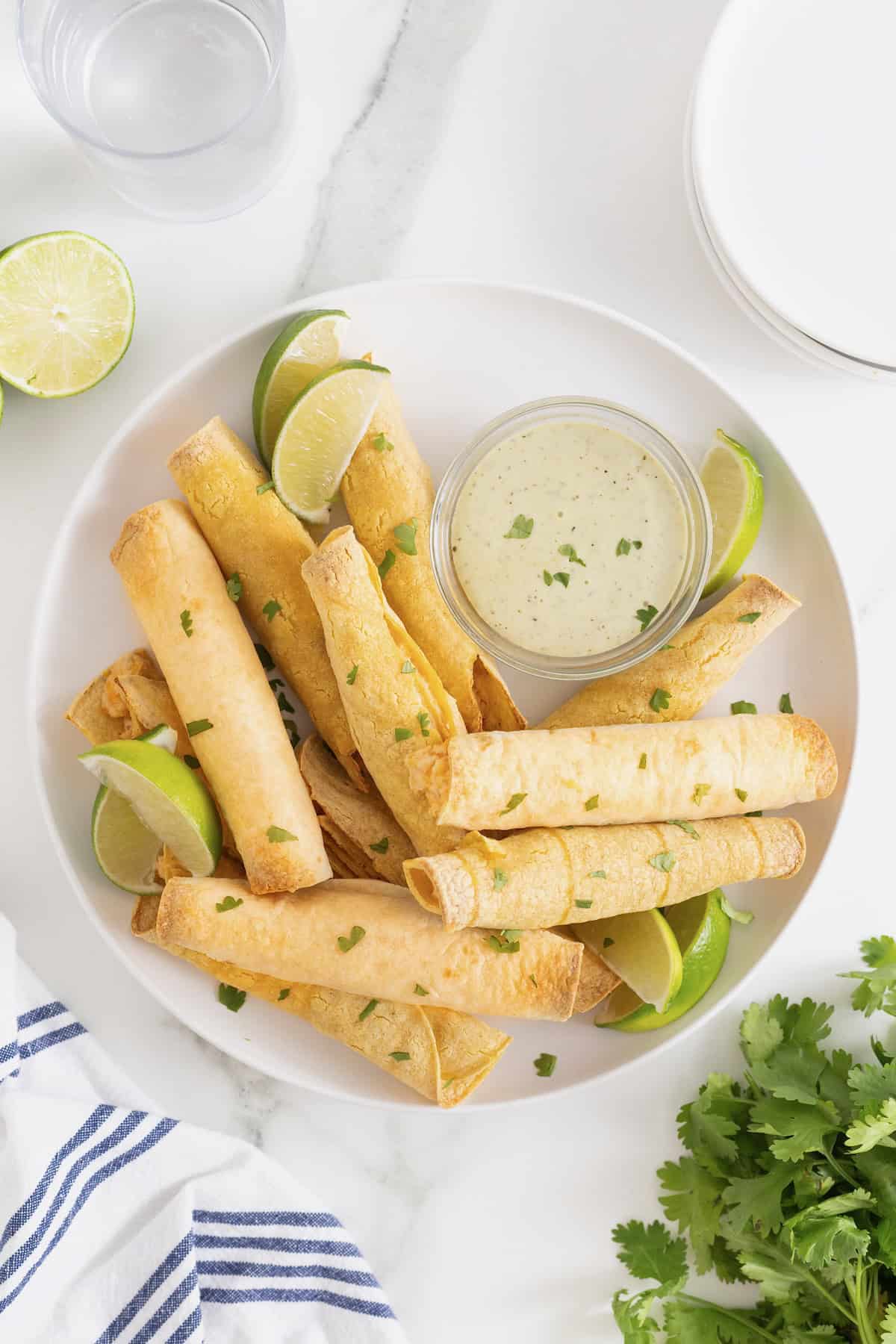 A pile of bacon cheddar ranch taquitos on a white plate with a small dish of ranch dressing.