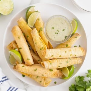 A pile of bacon cheddar ranch taquitos on a white plate with a small dish of ranch dressing.