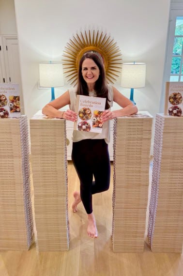 Maegan Brown standing next to four stacks of cookbooks, each up to her mid-chest. Maegan is holding a copy of her Celebration Boards cookbook.