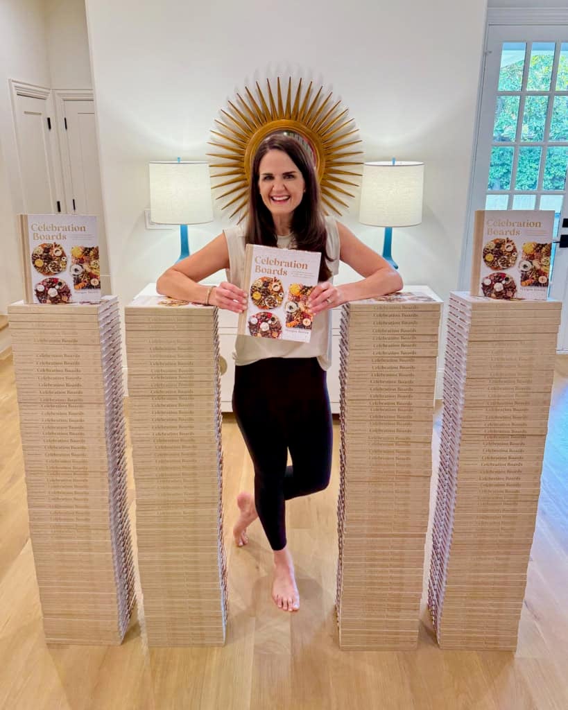 Maegan Brown standing next to four stacks of cookbooks, each up to her mid-chest. Maegan is holding a copy of her Celebration Boards cookbook.