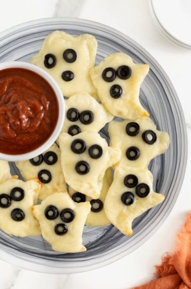 A gray plate piled with ghost shaped tortilla pizzas and a small white dish of marinara sauce.