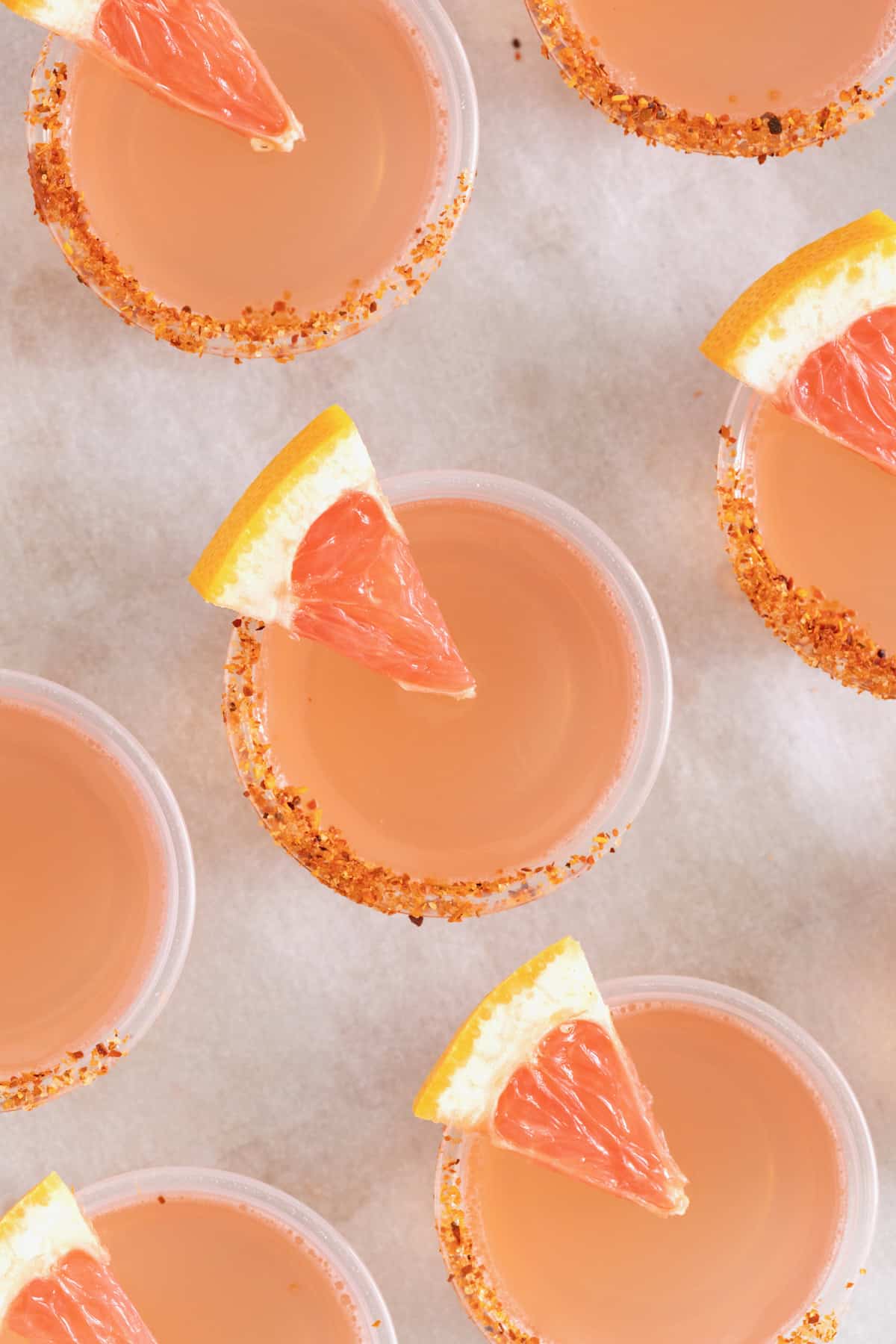 Pink colored jello shots in small plastic shot cups lined with Tajin and garnished with a small wedge of grapefruit.