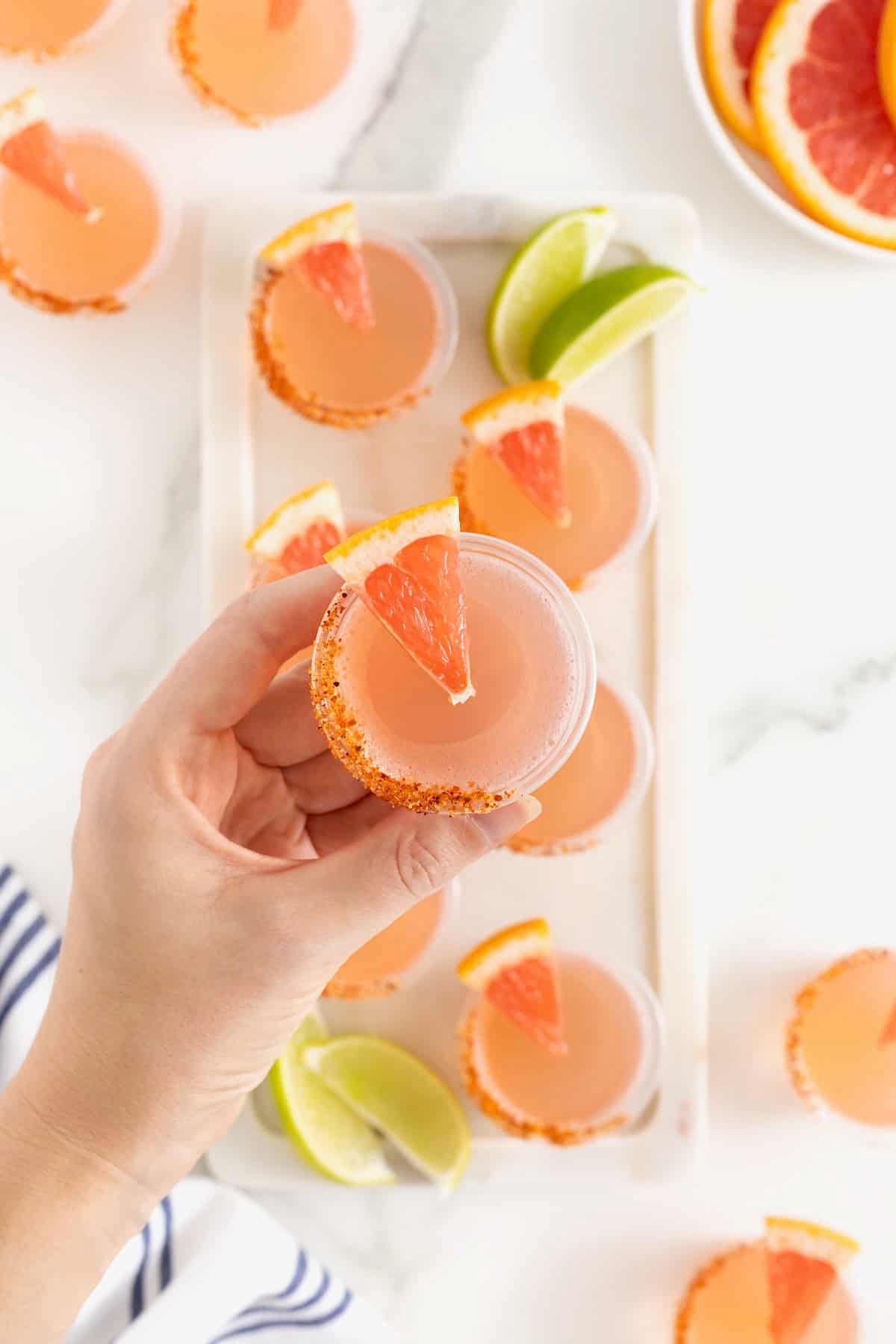 A hand holding a pink Paloma jello shot garnished with a TajÃn rim and a small wedge of grapefruit.