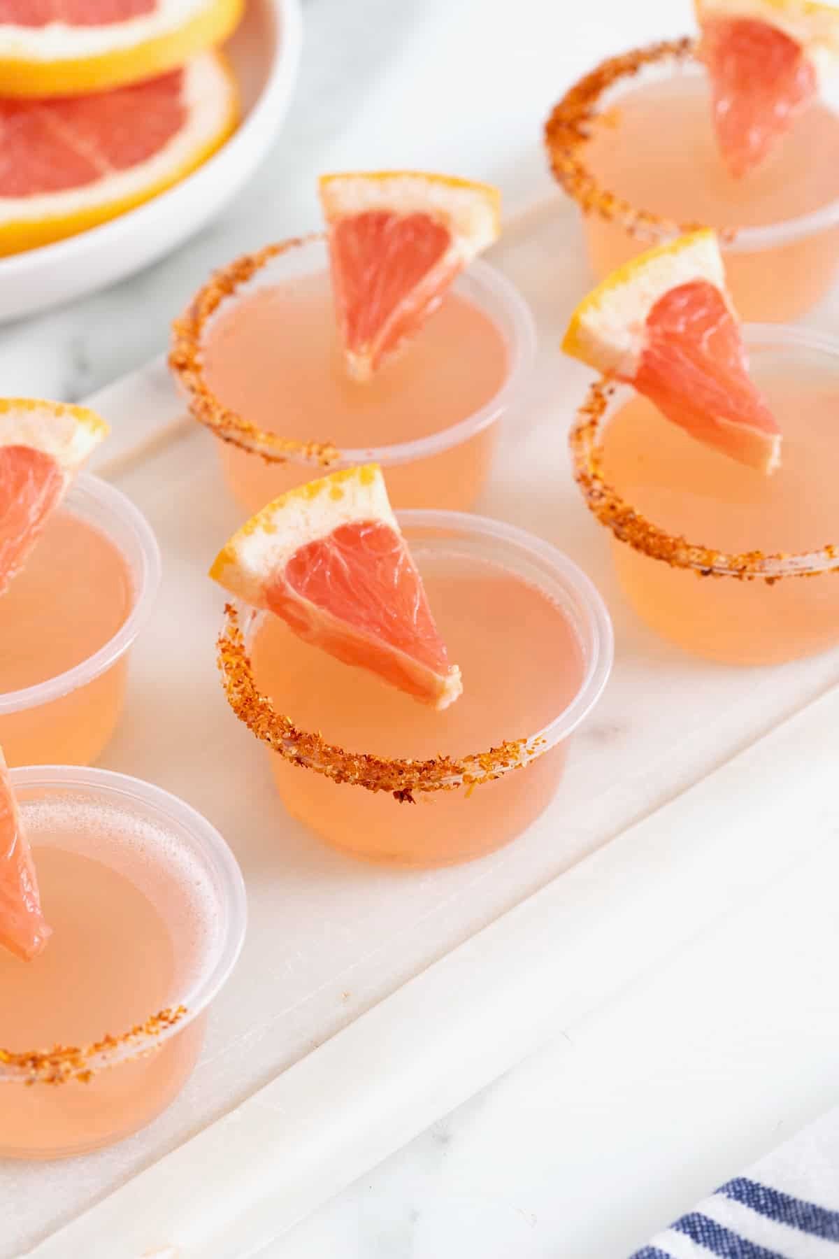 Pink colored jello shots in small plastic shot cups lined with Tajin and garnished with a small wedge of grapefruit.