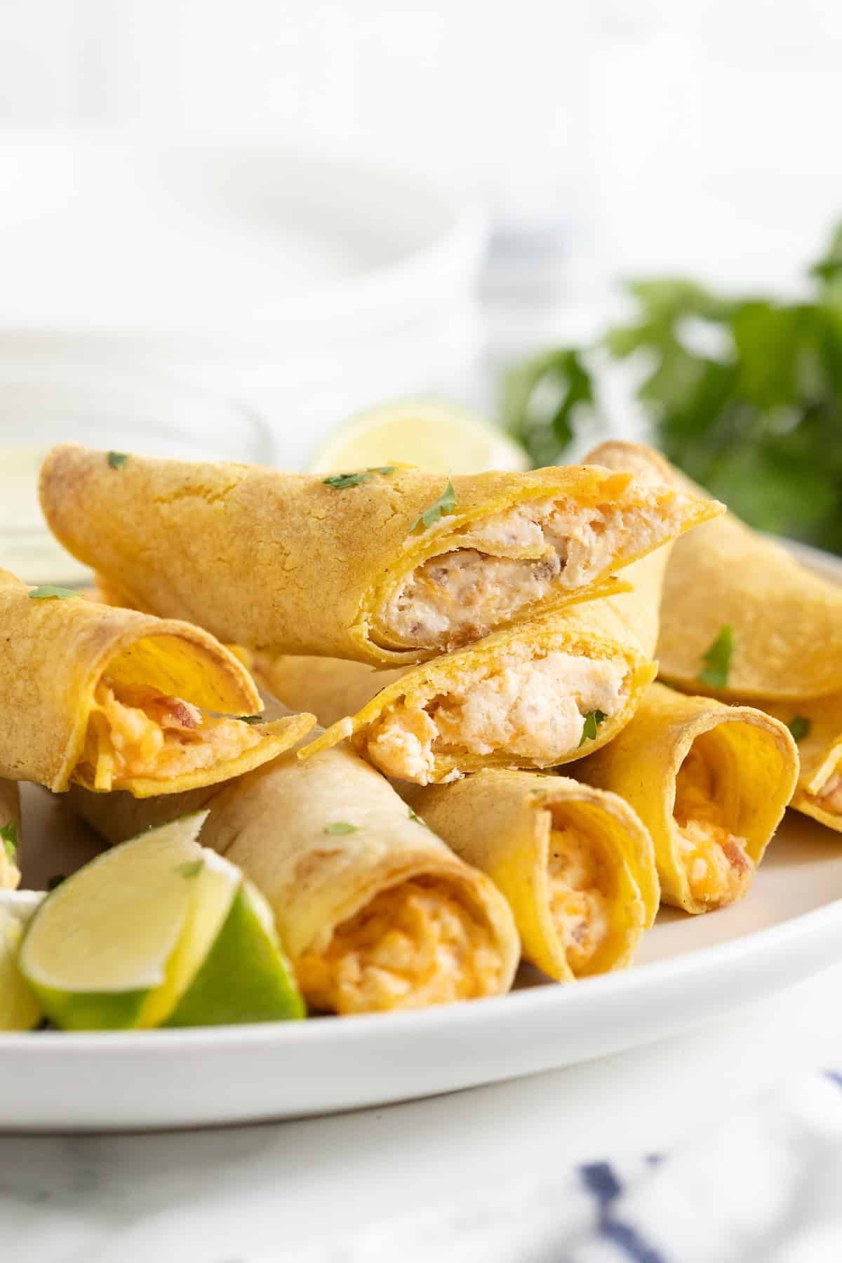 A pile of chicken bacon cheddar ranch taquitos on a white plate. Some of the taquitos are cut to reveal the filling.