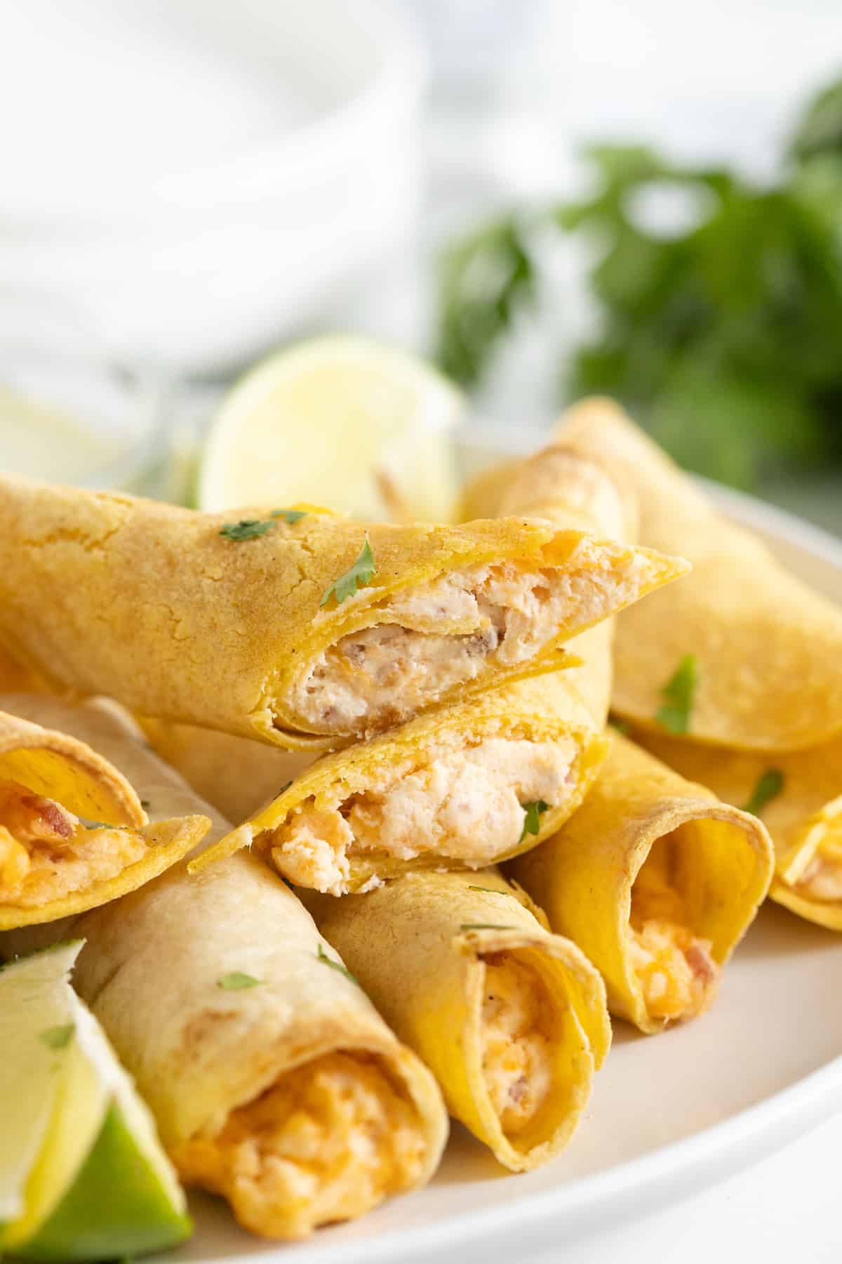 A pile of chicken bacon cheddar ranch taquitos on a white plate. Some of the taquitos are cut to reveal the filling.