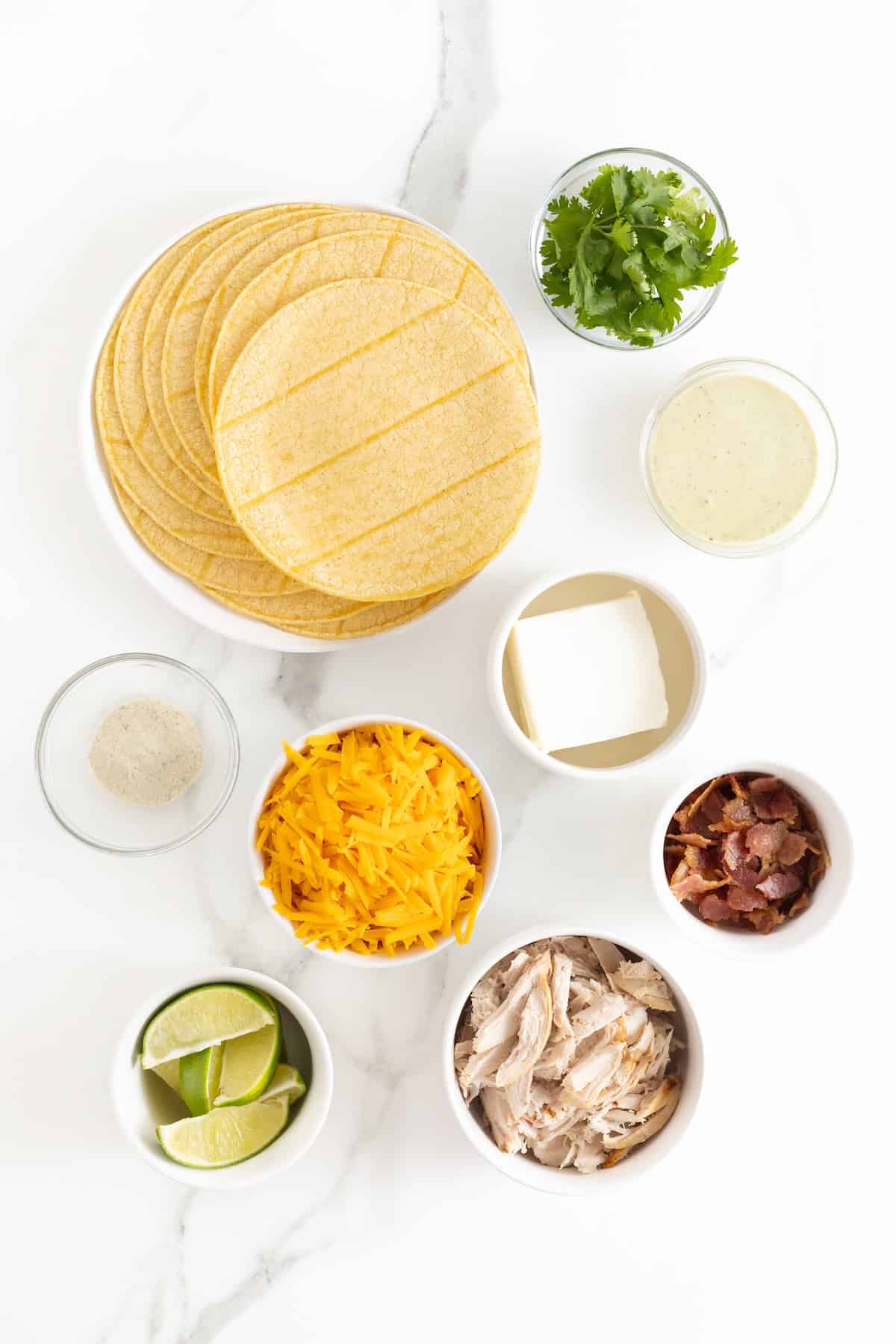 Ingredients to make bacon cheddar ranch chicken taquitos in small white dishes on a white marble counter.