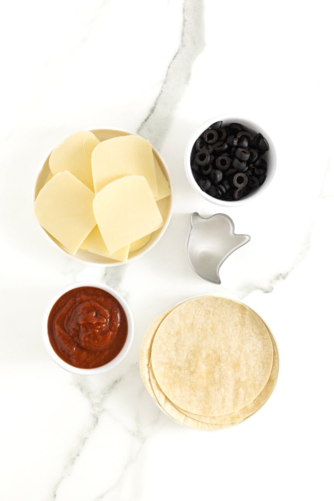 Ingredients to make ghost tortilla pizzas on a white marble counter.