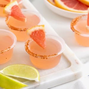 Pink colored jello shots in small plastic shot cups lined with Tajin and garnished with a small wedge of grapefruit.