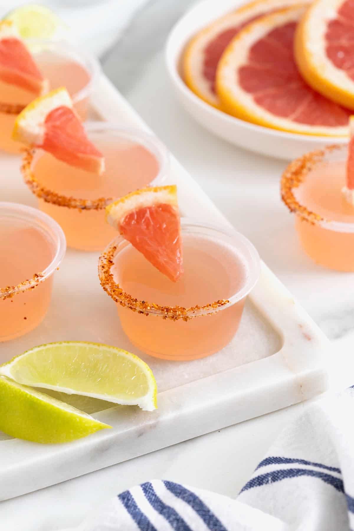 Pink colored jello shots in small plastic shot cups lined with pink salt and garnished with a small wedge of grapefruit.