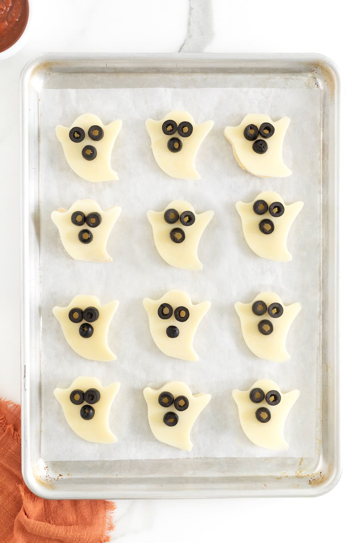 12 ghost shaped tortilla pizzas with sliced olives for eyes and mouths on a parchment lined baking sheet.