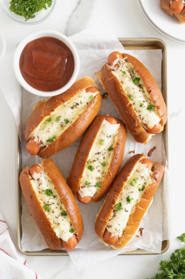 Six cheesy garlic bread hot dogs on a parchment lined aluminum baking sheet.