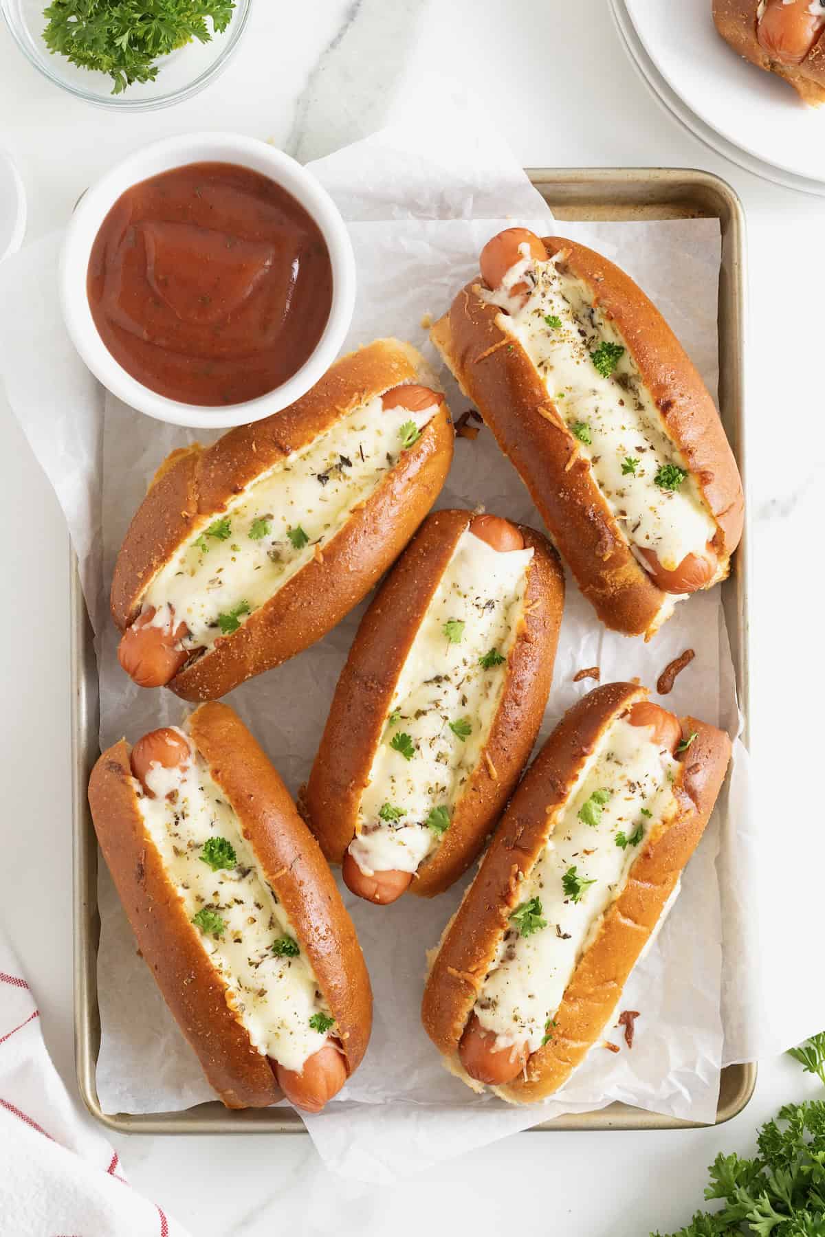 Six cheesy garlic bread hot dogs on a parchment lined aluminum baking sheet.