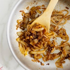 A light colored sauté pan with caramelized onions being stirred by a wooden spoon.