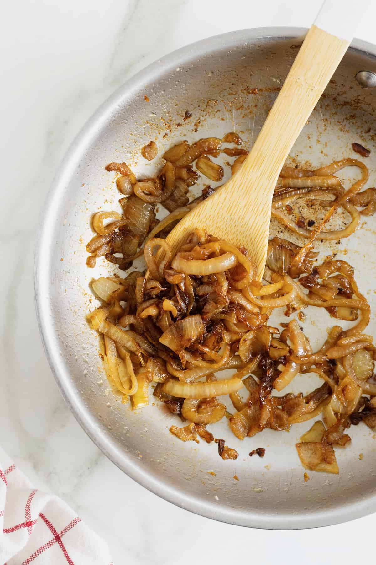 A light colored sauté pan with caramelized onions being stirred by a wooden spoon.