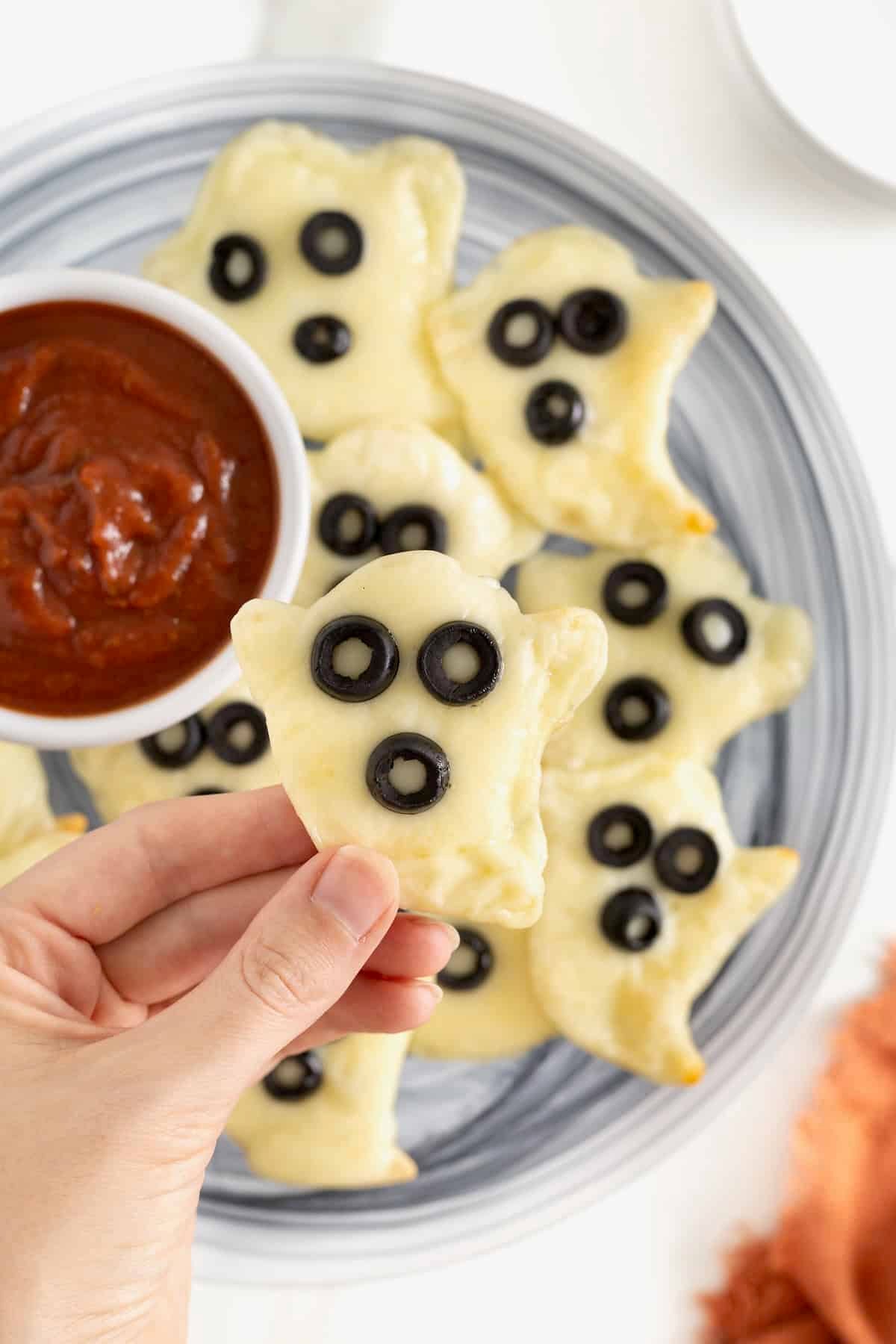 A gray plate piled with ghost shaped tortilla pizzas and a small white dish of marinara sauce.