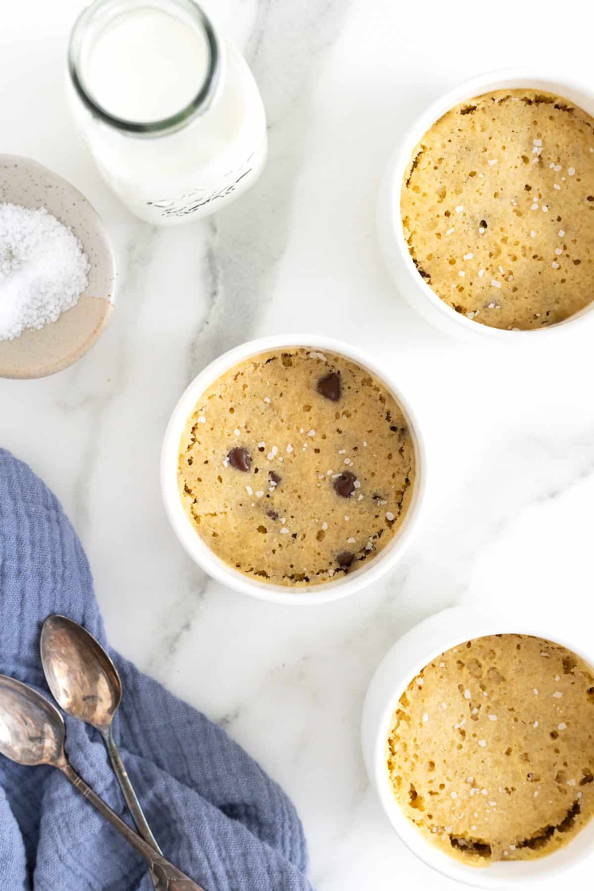 Three microwave chocolate chip cookies in white ramekins on a white marble counter.