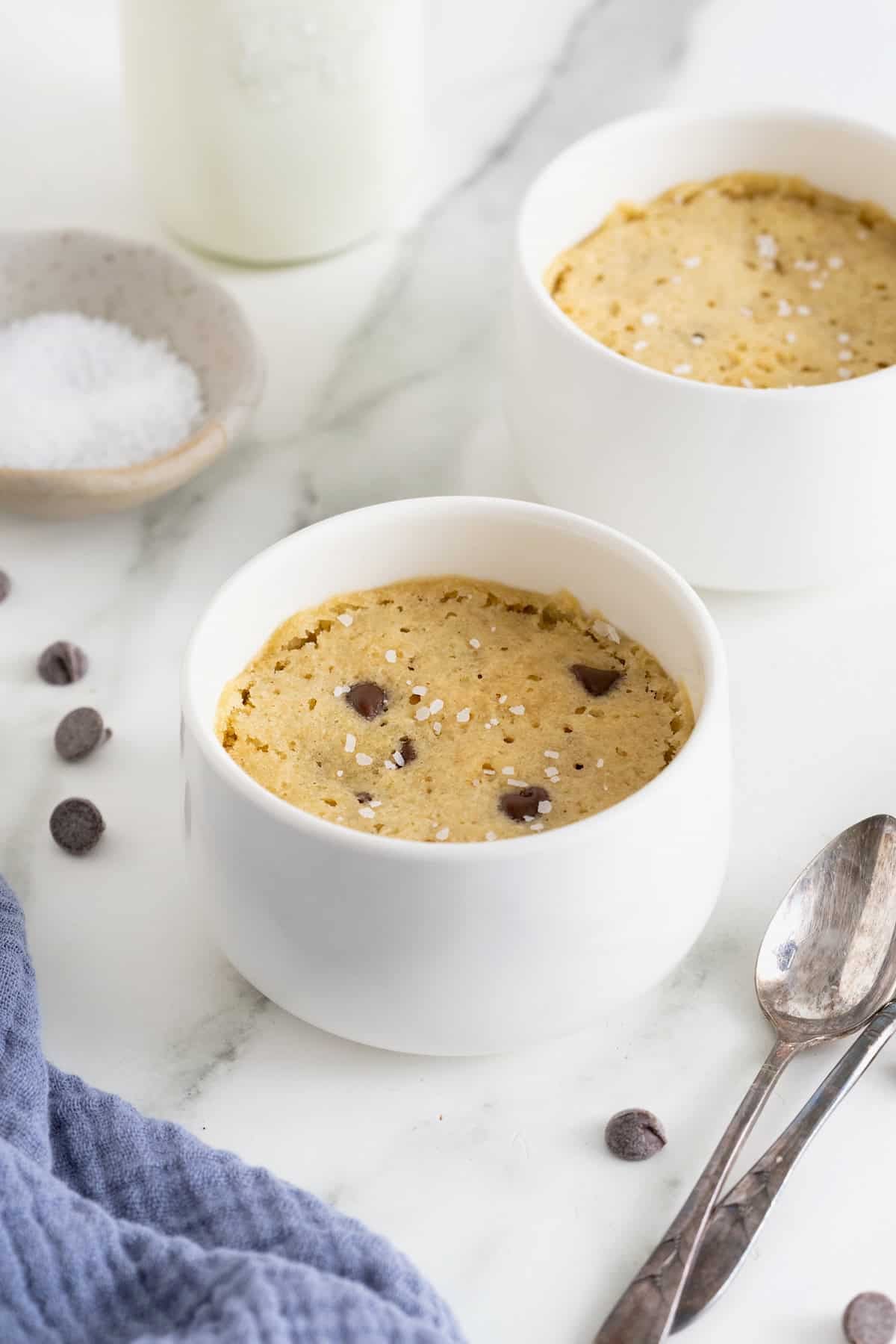 A microwave chocolate chip cookie in a white ramekin on a white marble counter next to two spoons.