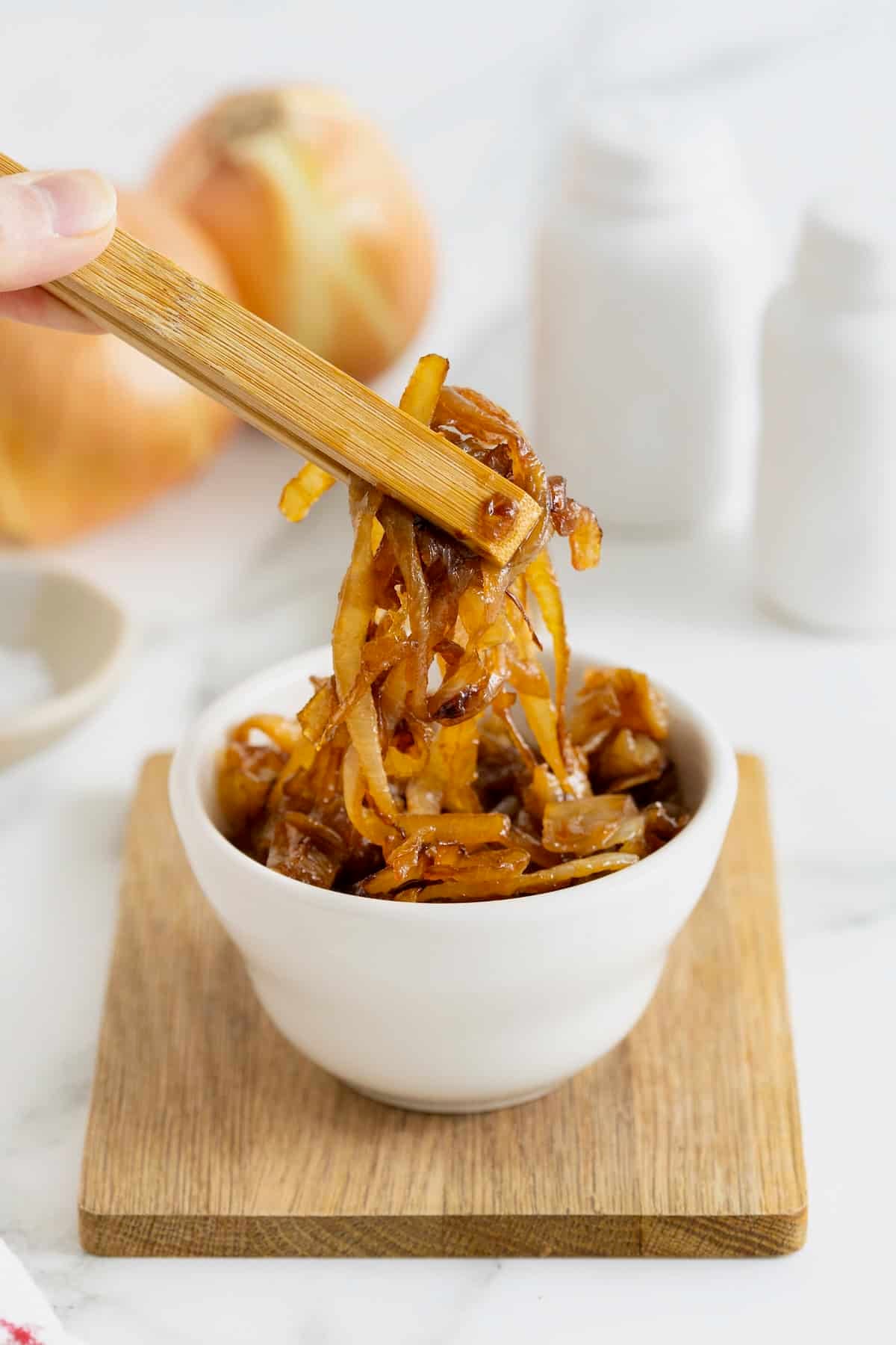 Wooden tongs lifting caramelized onions from a small white dish.