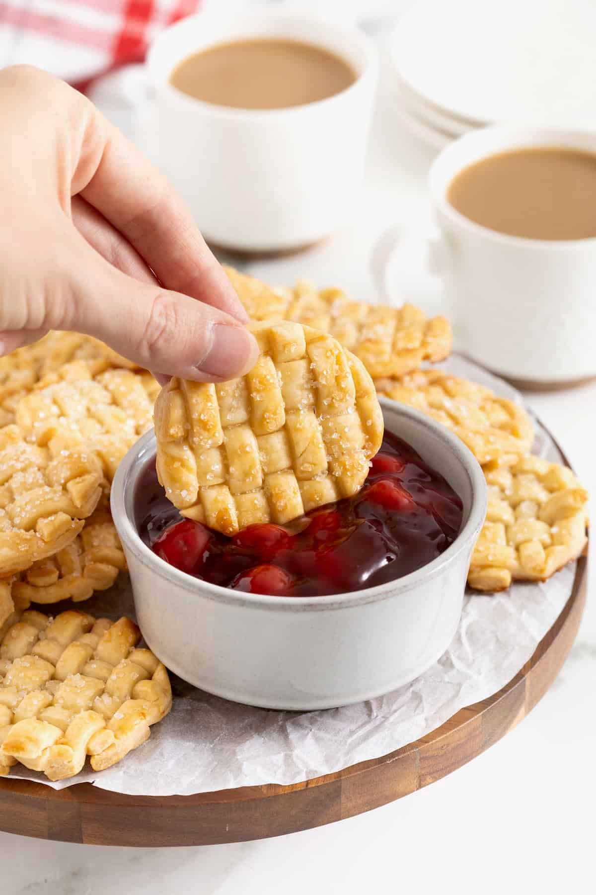A hand dipping a lattice pie cookie into a white ramekin of cherry pie filling.