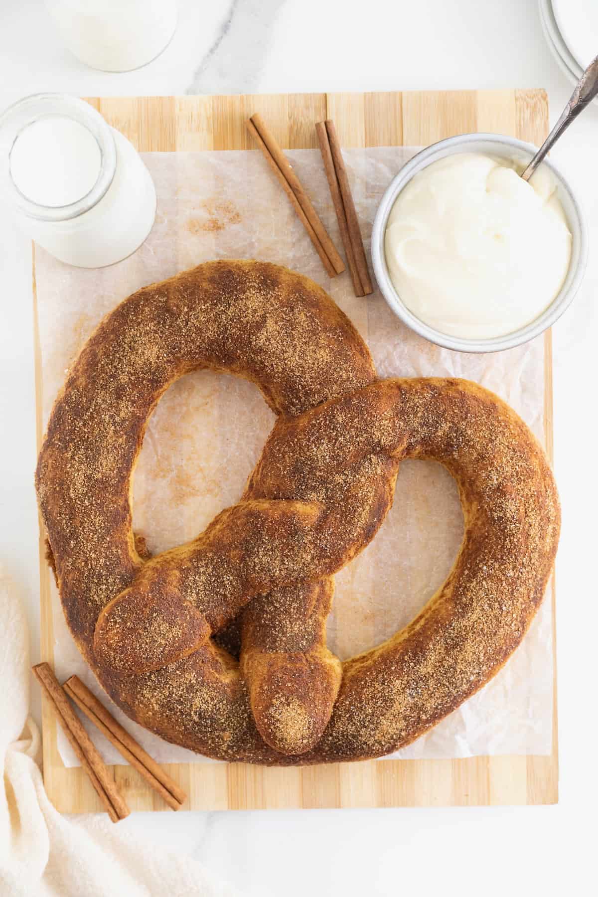 A jumbo sized soft pretzel covered in cinnamon sugar on a parchment lined wood cutting board. Next to the pretzel is a dish of cream cheese frosting and a few cinnamon sticks.